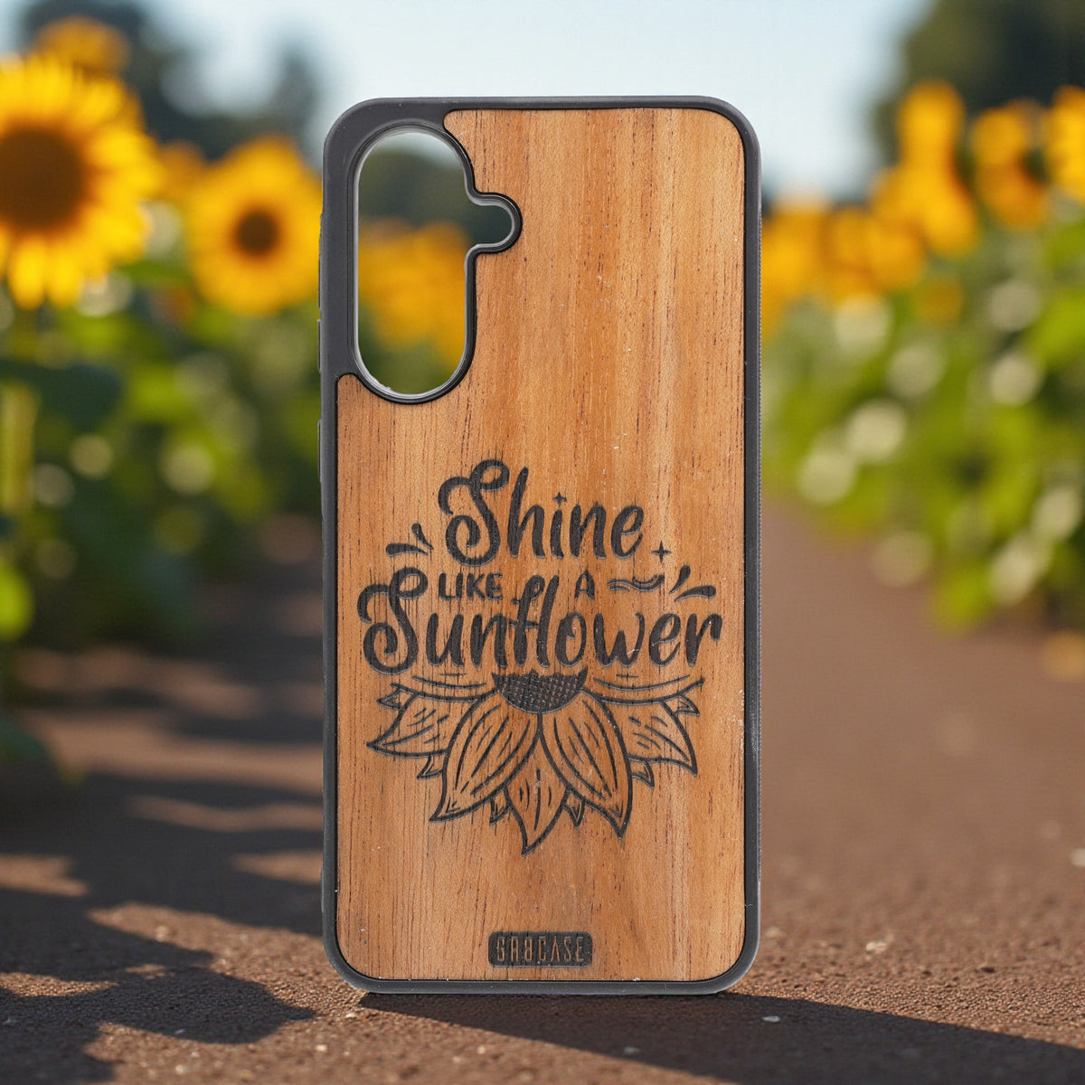 Wooden phone case with &#39;Shine Like a Sunflower&#39; design in front of sunflowers