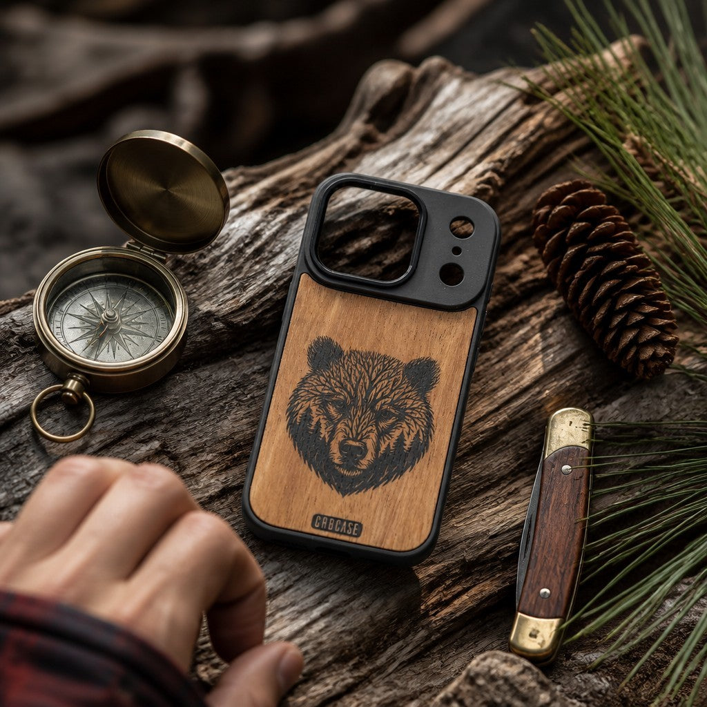 Hand-finished natural wood phone case featuring an intricate laser engraving of a grizzly bear head formed by tree branches and forest pines.